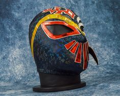 a colorful mask is displayed on a black stand against a blue background with red, yellow and orange accents