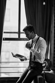 a man sitting on a window sill reading a book while wearing suspenders and a tie