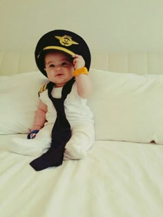 a baby wearing a hat and tie sitting on top of a white bed covered in pillows