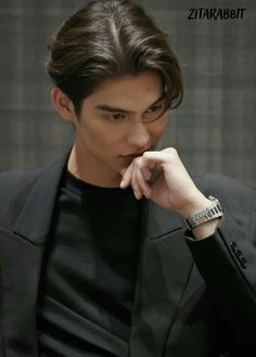 the young man is wearing a black suit and looking down at his hand on his chin