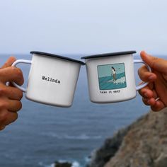 two people holding coffee mugs in front of the ocean with an image of a surfer on them