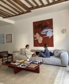 two people sitting on a couch in a living room next to a coffee table and large painting