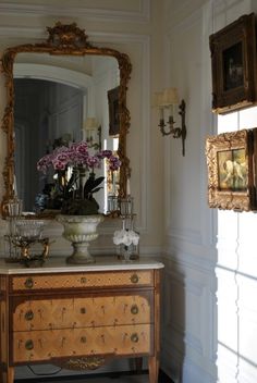 a dresser with flowers on it in front of a mirror and two framed pictures hanging on the wall