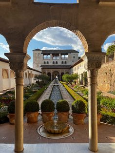 an outdoor fountain surrounded by potted plants in front of a large building with arches