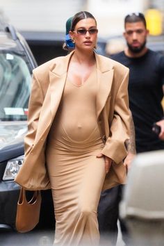 the pregnant woman is walking down the street in her tan dress and coat, carrying a brown handbag