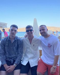 three men are posing for the camera while sitting on a bench by the beach with their arms around each other