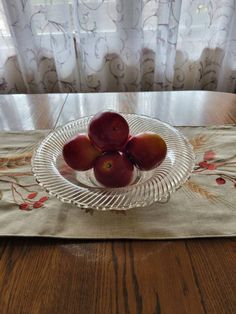 three plums in a glass bowl on a table with a window behind it and curtains