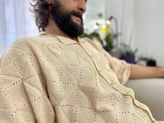 a bearded man sitting on a couch wearing a white knitted sweater and looking off to the side