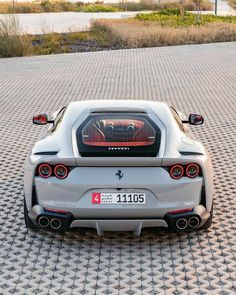 the rear end of a silver sports car parked on a cobblestone surface with its lights on