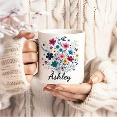 a woman is holding a coffee mug with flowers on it and the words ashley written in black