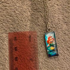 the little mermaid necklace has been placed next to a measuring ruler on a gray carpet