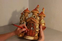 a hand holding a pencil near a clay model of a house with turrets on it