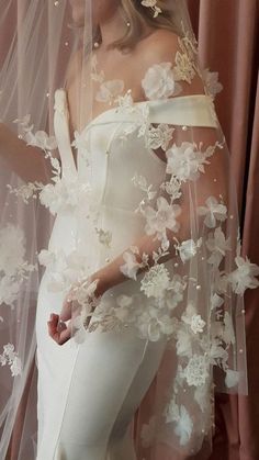 a woman wearing a white wedding gown and veil with flowers on the side, standing in front of a pink curtain