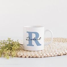 a white coffee mug with the letter r on it next to some dried flowers and a wicker place mat