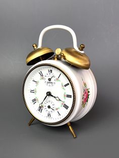 an alarm clock with flowers painted on the face and gold trimmings, sits in front of a gray background