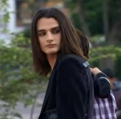 a young man with long hair walking down the street wearing a black jacket and jeans