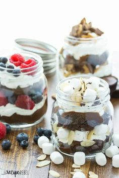 three mason jars filled with desserts on top of a wooden table next to berries and marshmallows