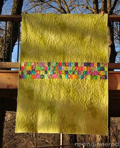 a yellow quilt hanging from a wooden fence with trees in the back ground and blue sky behind it