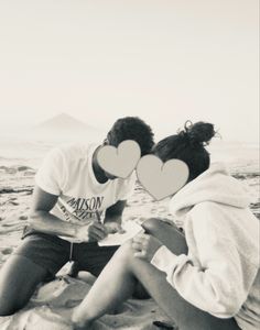 two people sitting in the sand with paper hearts on their faces and one person holding a cell phone