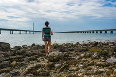 a woman standing on rocks near the ocean with a long bridge in the back ground