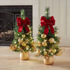 two small christmas trees with red bows and gold ornaments are on display in front of a fireplace