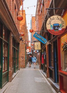 two people walking down an alleyway between buildings with signs on the sides and lights hanging above them