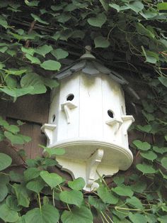 a white birdhouse hanging from the side of a building surrounded by green leaves and vines