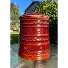 a red vase sitting on top of a table in front of a green bush and house