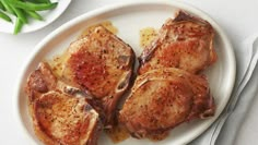 three pieces of meat on a white plate next to silverware and green beans in a bowl