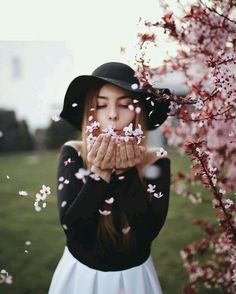 a woman in a black hat blowing white flowers on her face while standing next to a tree