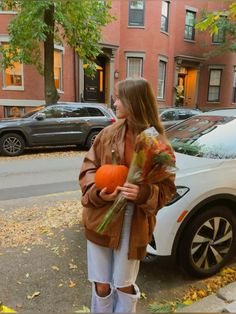 a young woman holding a pumpkin in front of a parked car on the side of a street