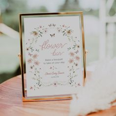 a flower bar sign sitting on top of a wooden table next to a white feather