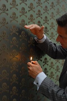 a man in a suit lighting a candle with skulls on the wall behind him,