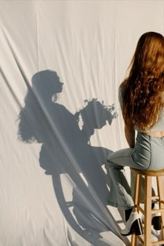 a woman sitting on top of a wooden stool next to a white sheet covered wall