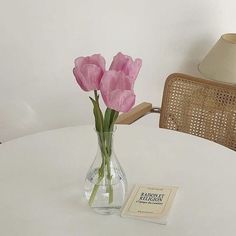 three pink flowers in a vase on a white table with a book and lamp next to it