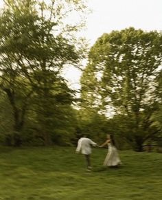 a man and woman holding hands walking through the grass with trees in the back ground