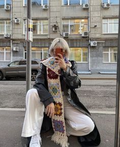 a woman sitting on the curb taking a selfie with her cell phone while wearing a scarf