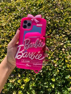 a person holding up a pink phone case with the words barbie on it in front of some bushes