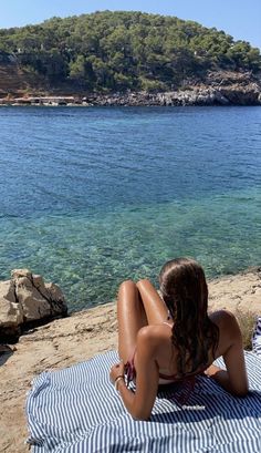 a woman sitting on top of a towel next to the ocean with an island in the background