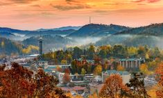 the city is surrounded by mountains and trees with fall foliage on them, as the sun sets in the distance