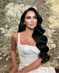 a woman with long black hair wearing a white dress in front of a floral wall