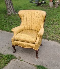 an old yellow chair sitting on the sidewalk in front of a tree and grass area