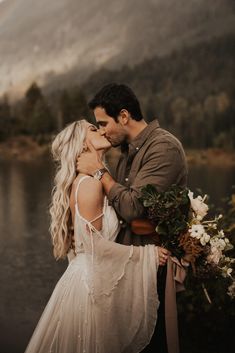 a bride and groom kissing in front of a mountain lake with flowers on the bouquet