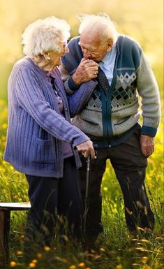 an older couple standing next to each other in a field with grass and yellow flowers