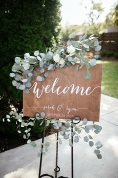 a welcome sign with greenery on it sitting on top of a metal stand in front of a tree