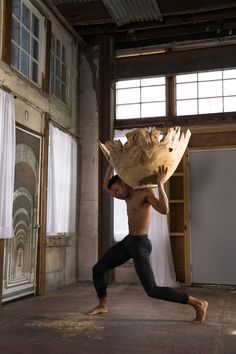 a shirtless man holding a large piece of wood in an empty room with windows