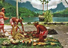 two men are preparing food on the ground in front of some water and mountains with palm trees