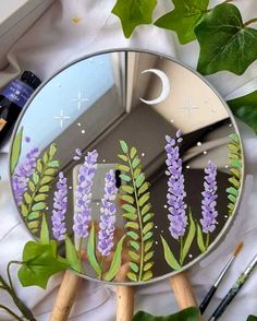 a mirror with flowers painted on it next to some paintbrushes and greenery