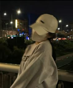 a woman wearing a white hat is standing on a balcony at night with city lights in the background