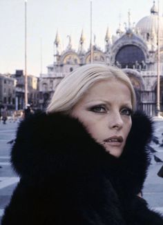 a woman with blonde hair wearing a black fur coat and looking at the camera, in front of an ornate building